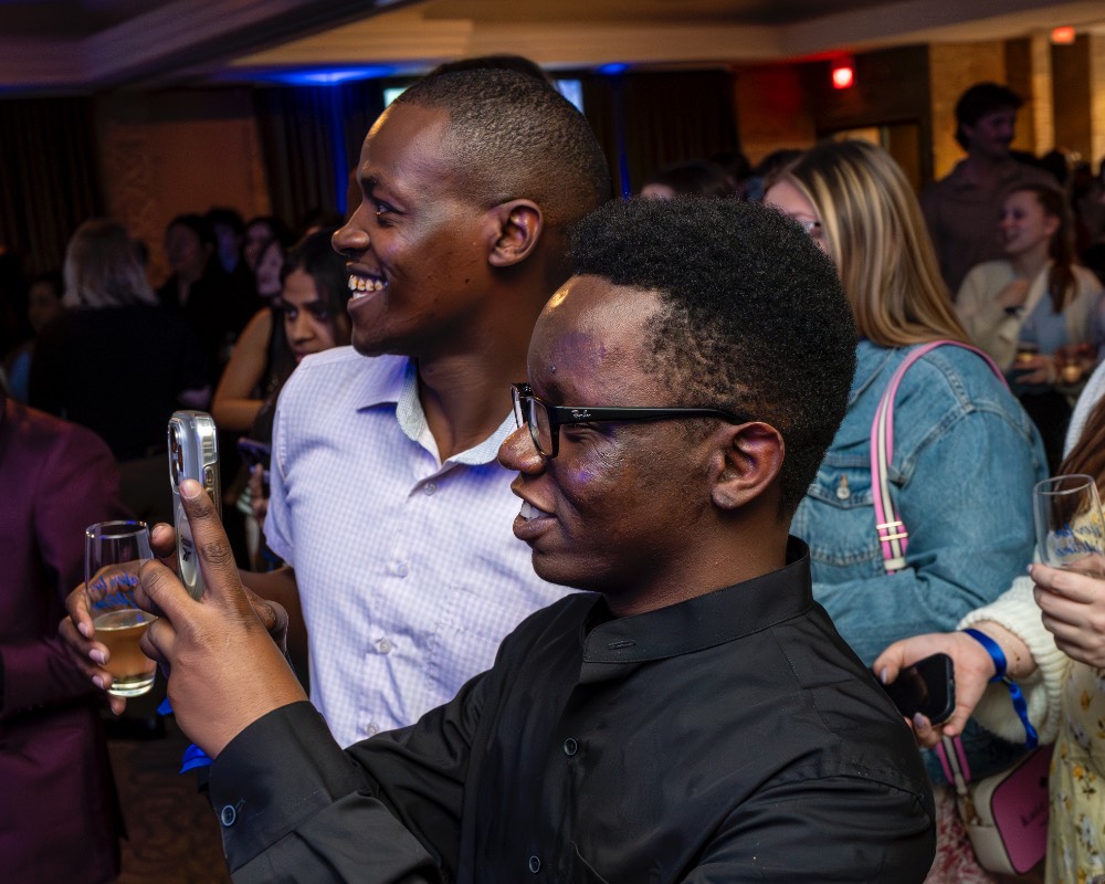 Two boys side by side, one hold champagne glass and the other holding phone.